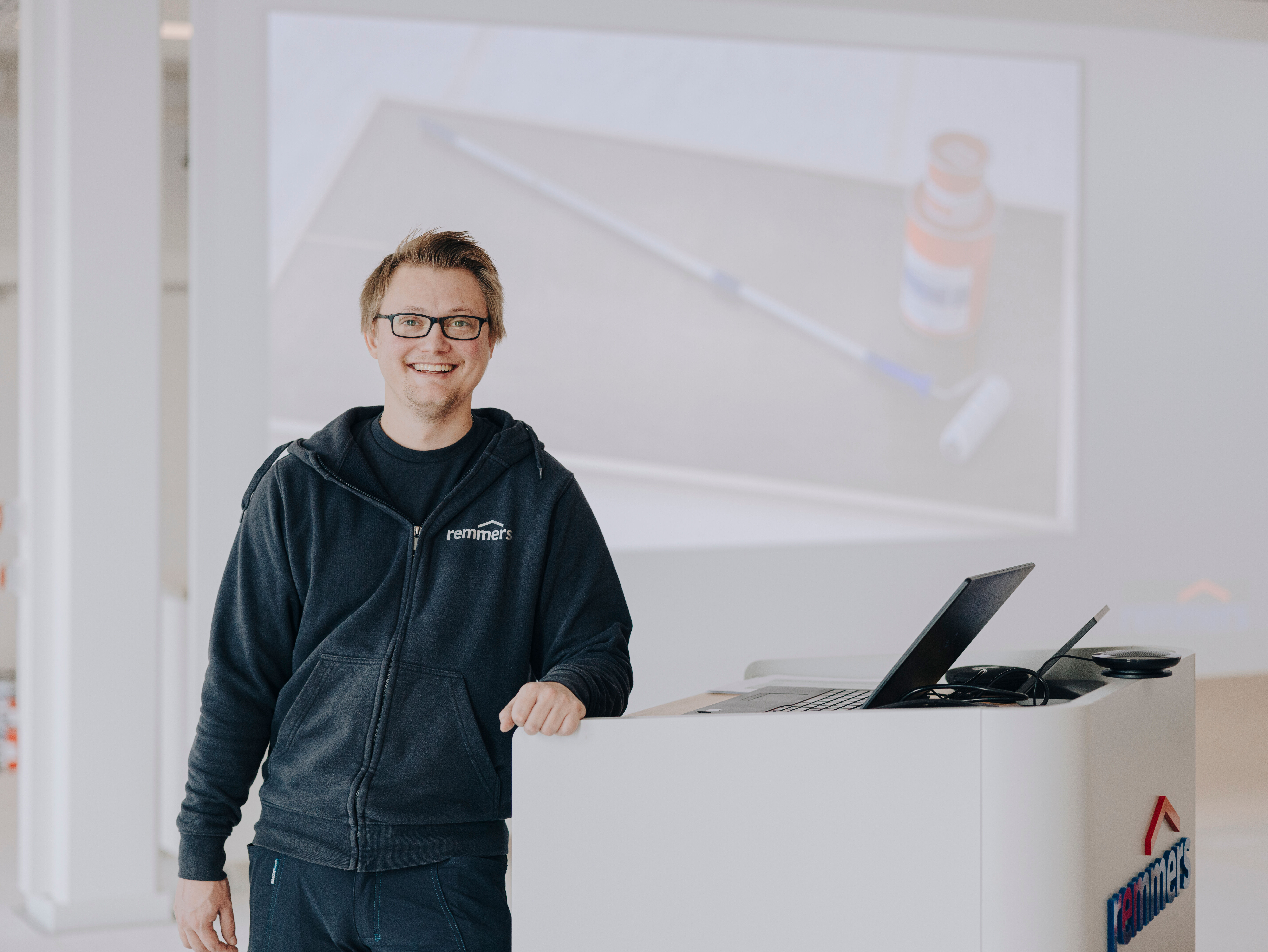 Person lehnt an Empfangstresen mit Laptop, Präsentationsfolie im Hintergrund zeigt Produkt und Anwendung im Innenraum.