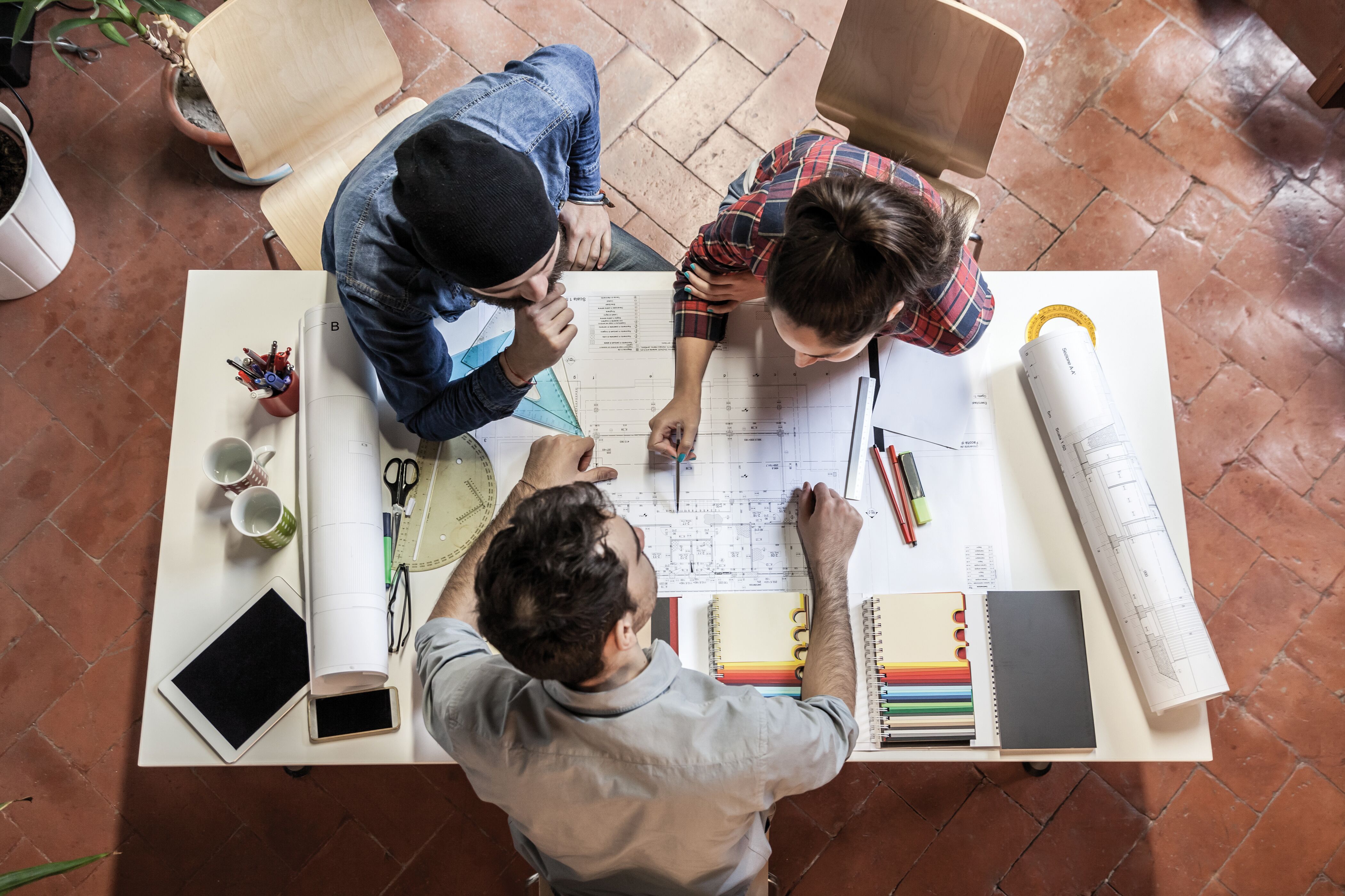 Draufsicht auf drei Personen am Tisch, arbeiten gemeinsam an Bauplan mit Zeichnungen, Stiften und Materialmustern.